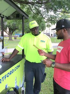 An African American man in bright lime green shirt and hat that matches his information cart discusses information on a cardboard flyer with an African-American man wearing a cap and red T-shirt. 